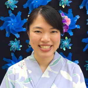 Headshot of Risa Katori. Risa is wearing a light blue yukata and standing in front of a black background with blue flowers.